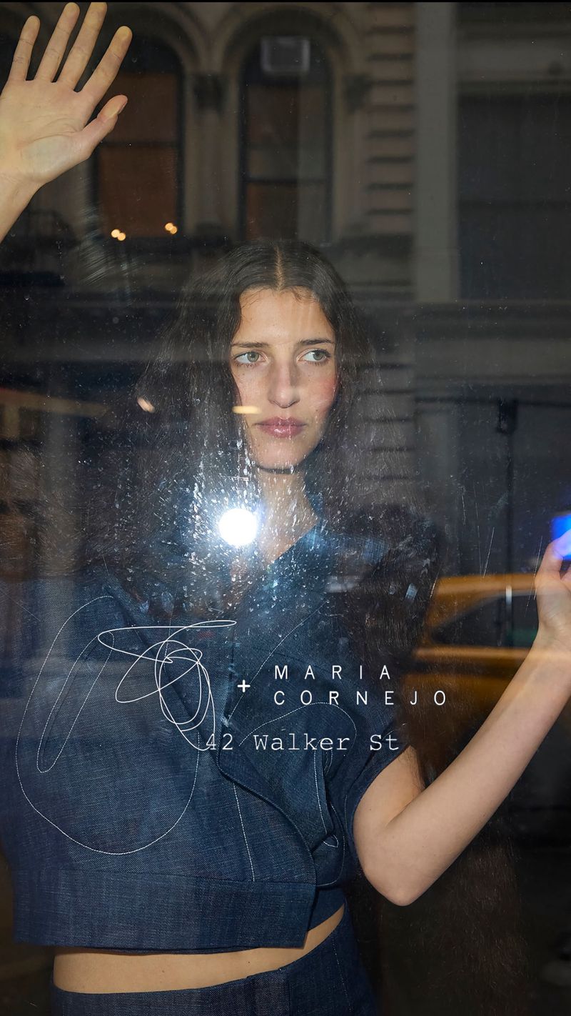 Indoor portrait of a woman in a blue denim outfit, posing behind a window with writing, against an urban building backdrop.
