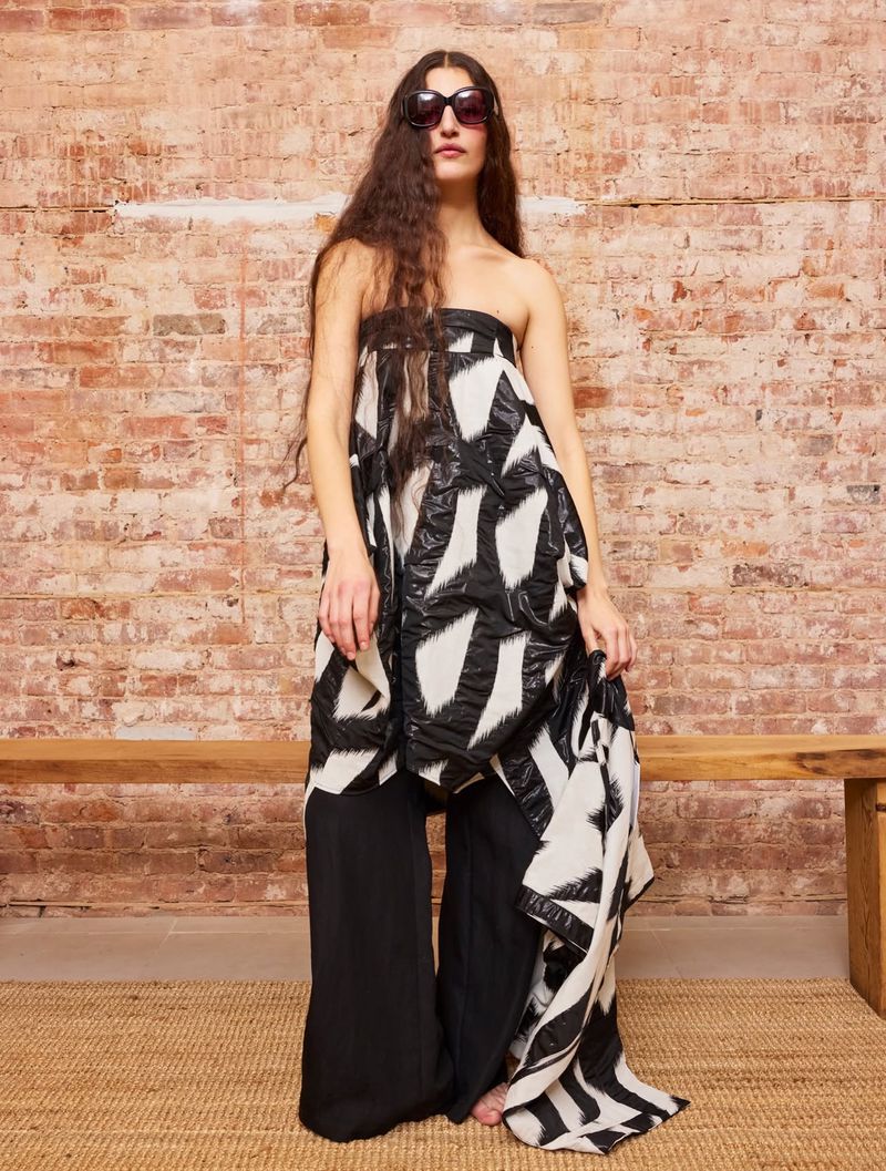 Model wearing dark sunglasses and a black and white patterned strapless dress against a brick wall background