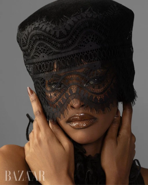 Close-up fashion shot of a woman with brown skin wearing a black lace mask and head covering, accented with glossy brown lips.