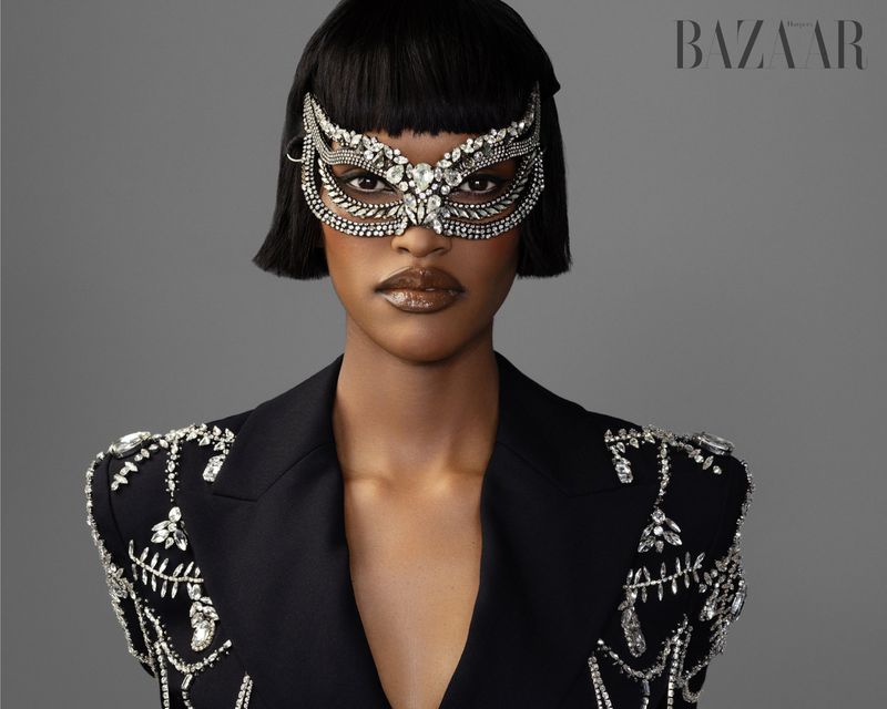Glamorous portrait of a black woman in a masquerade mask and black blazer with crystal embellishments, against a grey backdrop.