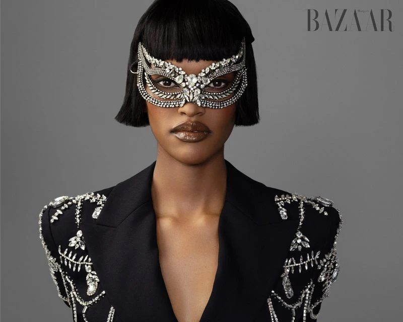 Glamorous portrait of a black woman in a masquerade mask and black blazer with crystal embellishments, against a grey backdrop.