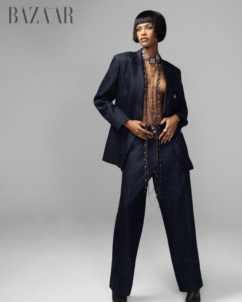 Fashion editorial shot of woman in a navy-blue suit with sheer embellished top on a grey background, with the Bazaar logo.