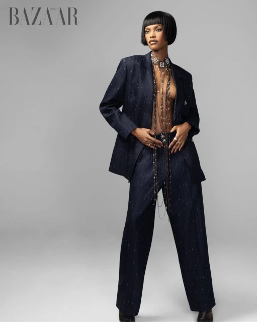 Fashion editorial shot of woman in a navy-blue suit with sheer embellished top on a grey background, with the Bazaar logo.