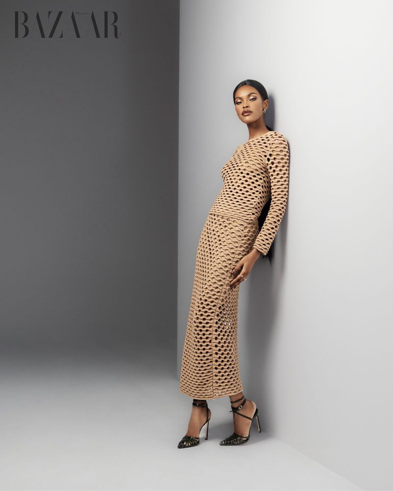Elegant woman in a tan, open-weave long-sleeved dress and high heels leans against a corner in a fashion editorial shot for Bazaar.