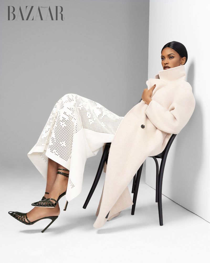 Elegant woman in stylish coat and heels seated on chair in studio against gray backdrop; high-fashion aesthetic, modern editorial.
