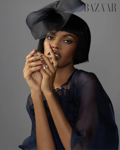Glamorous beauty shot of a woman in a sheer navy top and large navy hat, hands near her face showcasing gold rings and manicure, gray background.