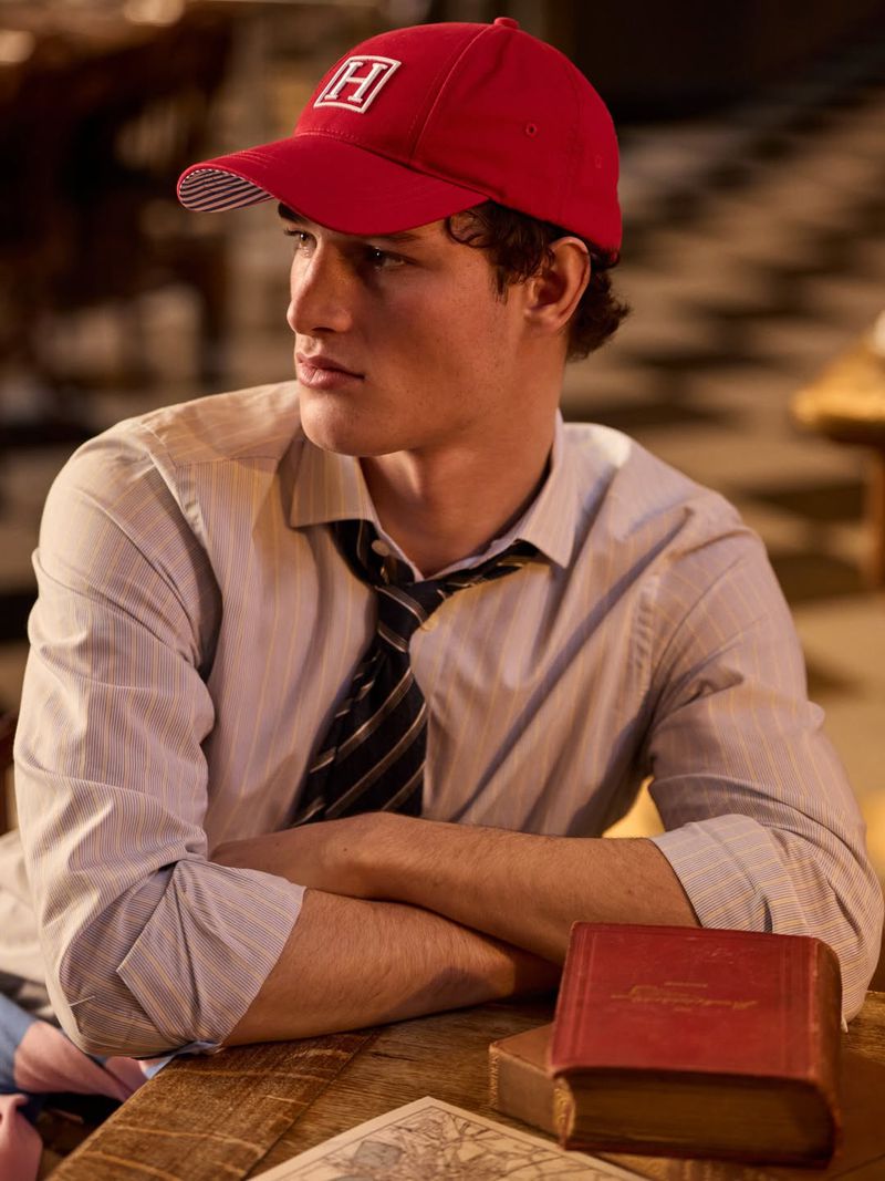 Young man wearing a red baseball cap and striped shirt seated at a wooden table with a book for Hackett Heritage Collection S/S 26
