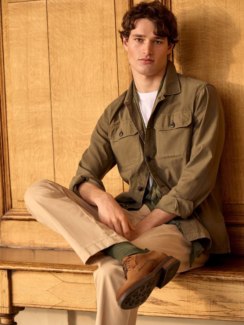 A young man seated in an olive green utility shirt and khaki trousers against a wooden paneling background.
