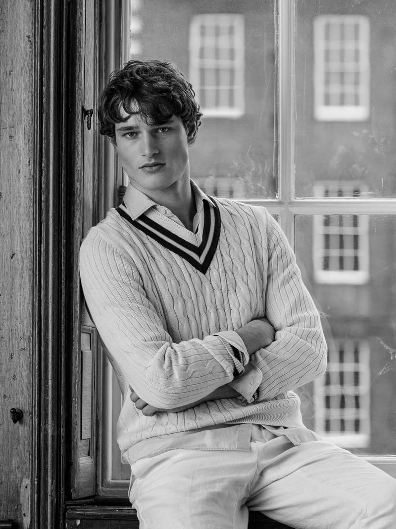 Portrait of male model Martin Antech wearing a cable-knit cricket sweater seated by a large window for Hackett
