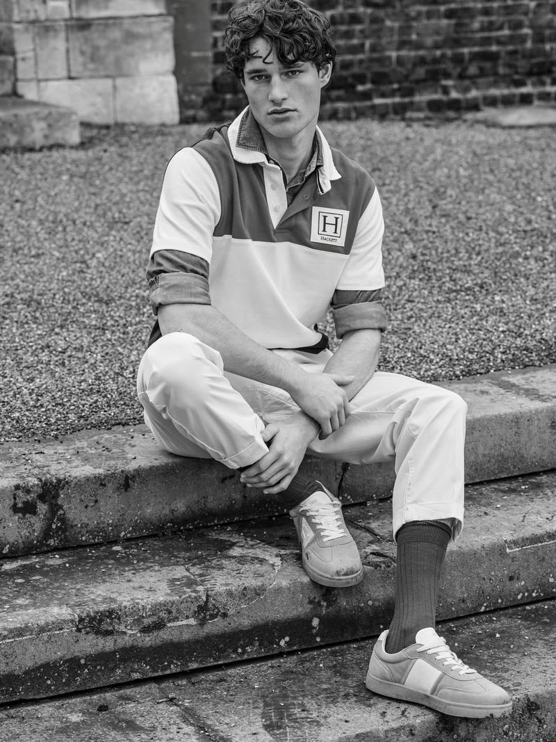 Model seated on stone steps wearing a colorblocked rugby shirt and light trousers for Hackett Heritage Collection