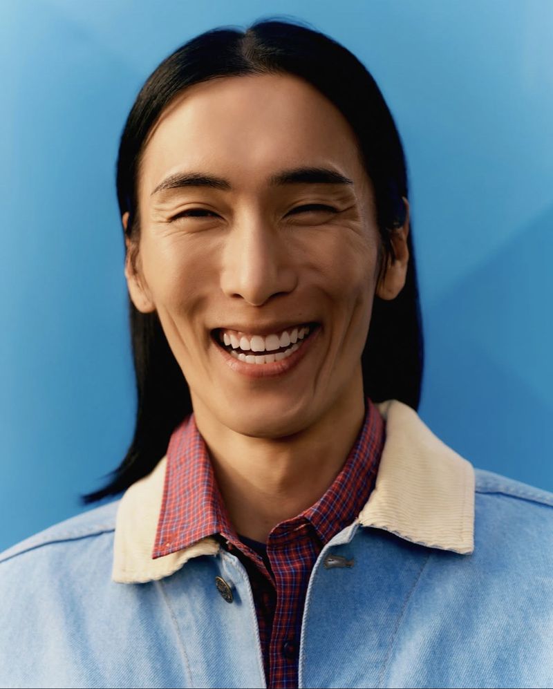 Close-up portrait of model Hyuk Kim smiling, wearing a blue jacket with a white collar over a red patterned shirt against a blue background.