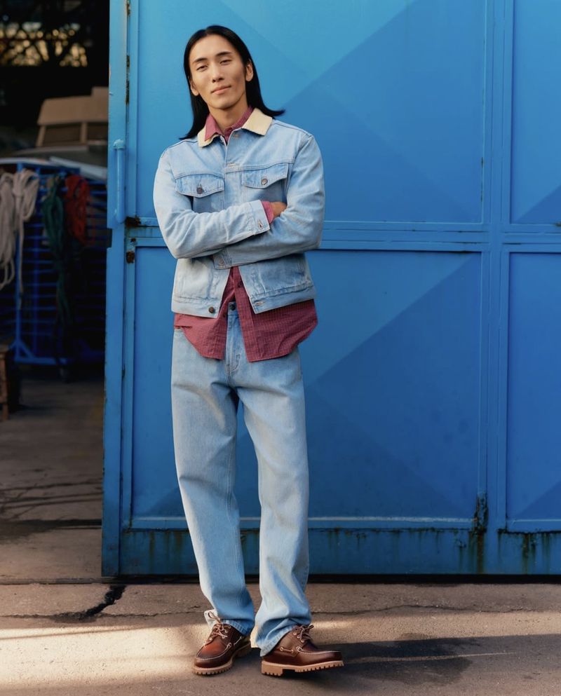 Model with long dark hair wearing a light denim jacket and brown boat shoes standing against a blue door.