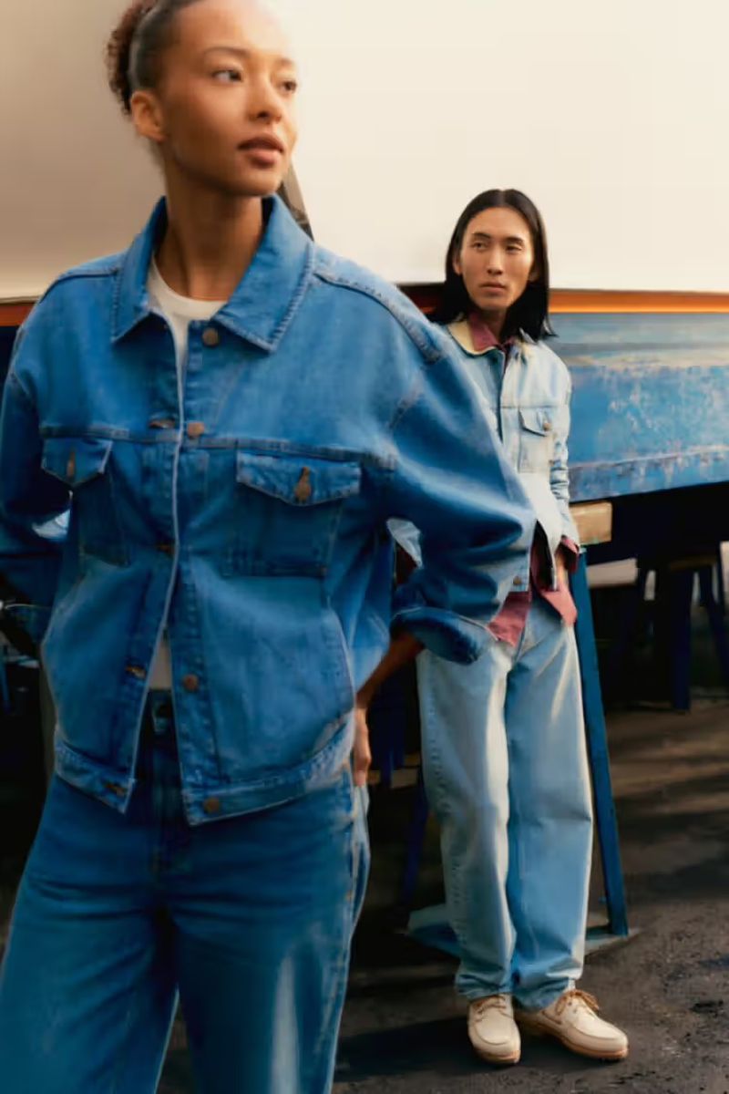 Two people in denim jackets and jeans pose in front of a partially visible boat. The light is diffused and the colors are muted.