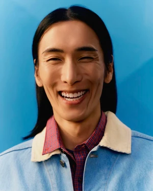 Close-up portrait of model Hyuk Kim smiling, wearing a blue jacket with a white collar over a red patterned shirt against a blue background.