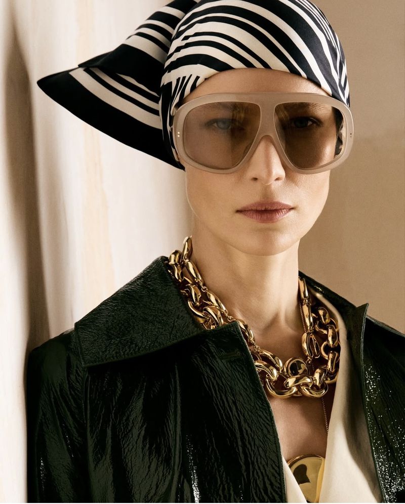Model wearing a black and white striped headscarf, translucent sunglasses, chunky gold chain necklace, and dark green coat.