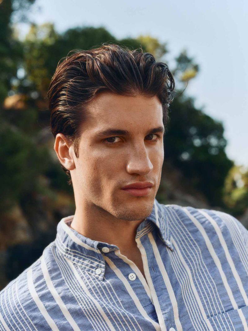 Close-up of a handsome man with dark hair wearing a blue and white striped button-down shirt, posing outdoors with a blurred background.