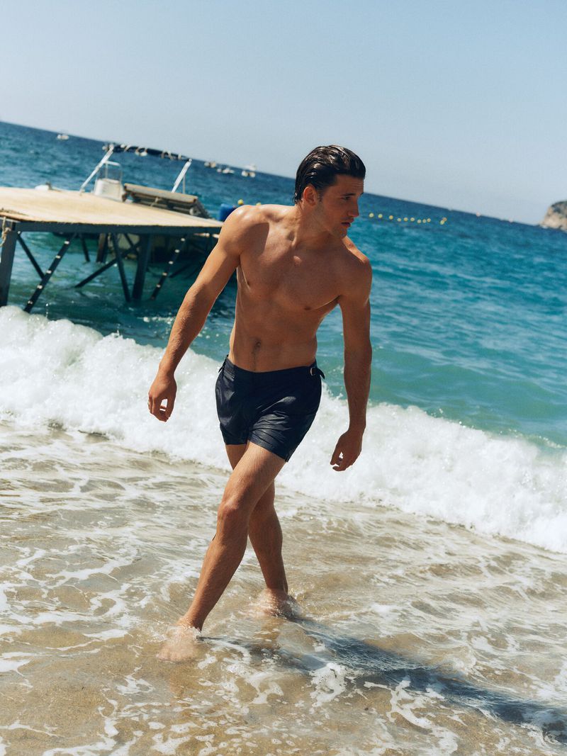 Shirtless male model walking through shallow ocean surf near a wooden dock under clear sky