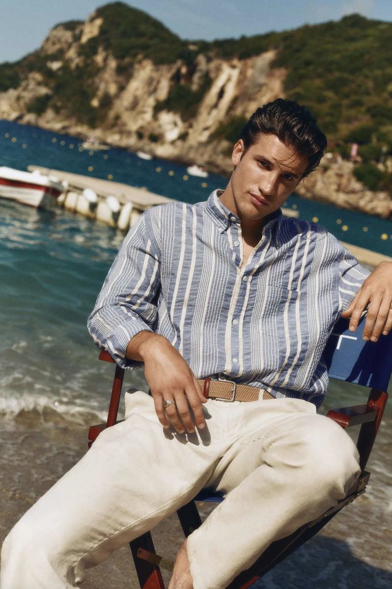 Man wearing a striped shirt and cream trousers seated on a director chair by the coast for Gant By The Coast project