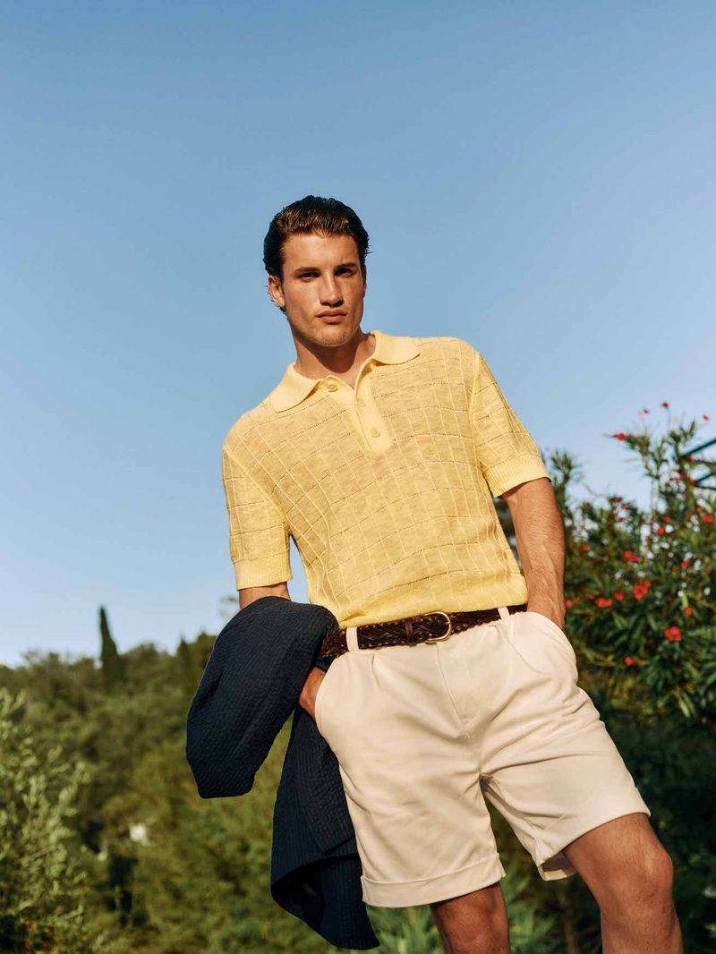 Man wearing a yellow knit polo shirt and white shorts holding a dark sweater against a clear sky background