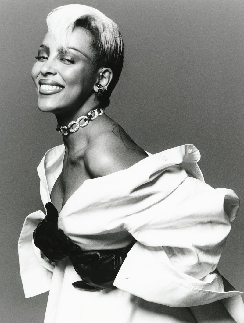 Elegant woman with platinum blonde hair and a choker poses in an unbuttoned white shirt and black gloves against a grey background.