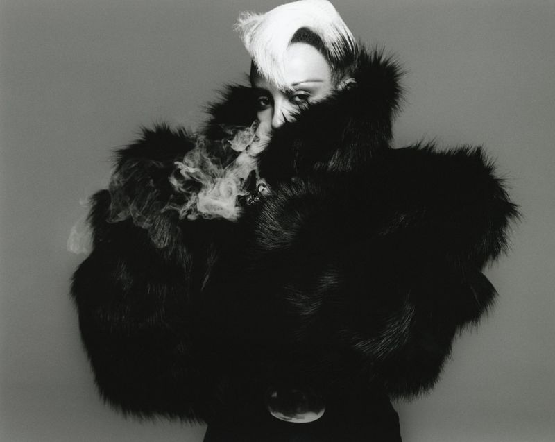 Striking black and white studio portrait of an adult female wearing a voluminous fur coat and elaborate platinum hairstyle.