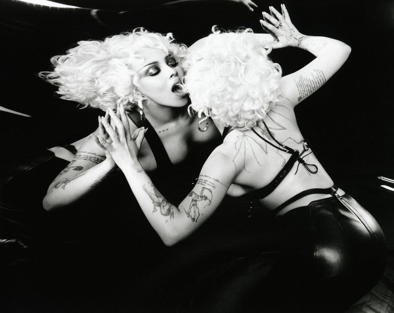 Monochrome fashion shot featuring an adult female with a blonde curly hairstyle, tattoos, and jewelry in a studio environment.