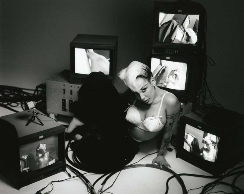 Young woman in bra poses amid vintage TV sets displaying various video clips in a monochrome-themed studio setting.