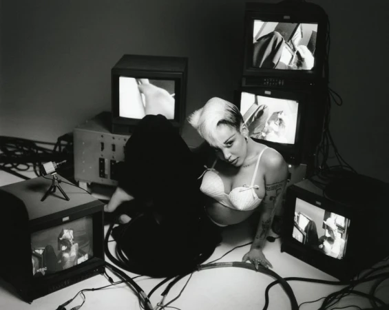 Young woman in bra poses amid vintage TV sets displaying various video clips in a monochrome-themed studio setting.
