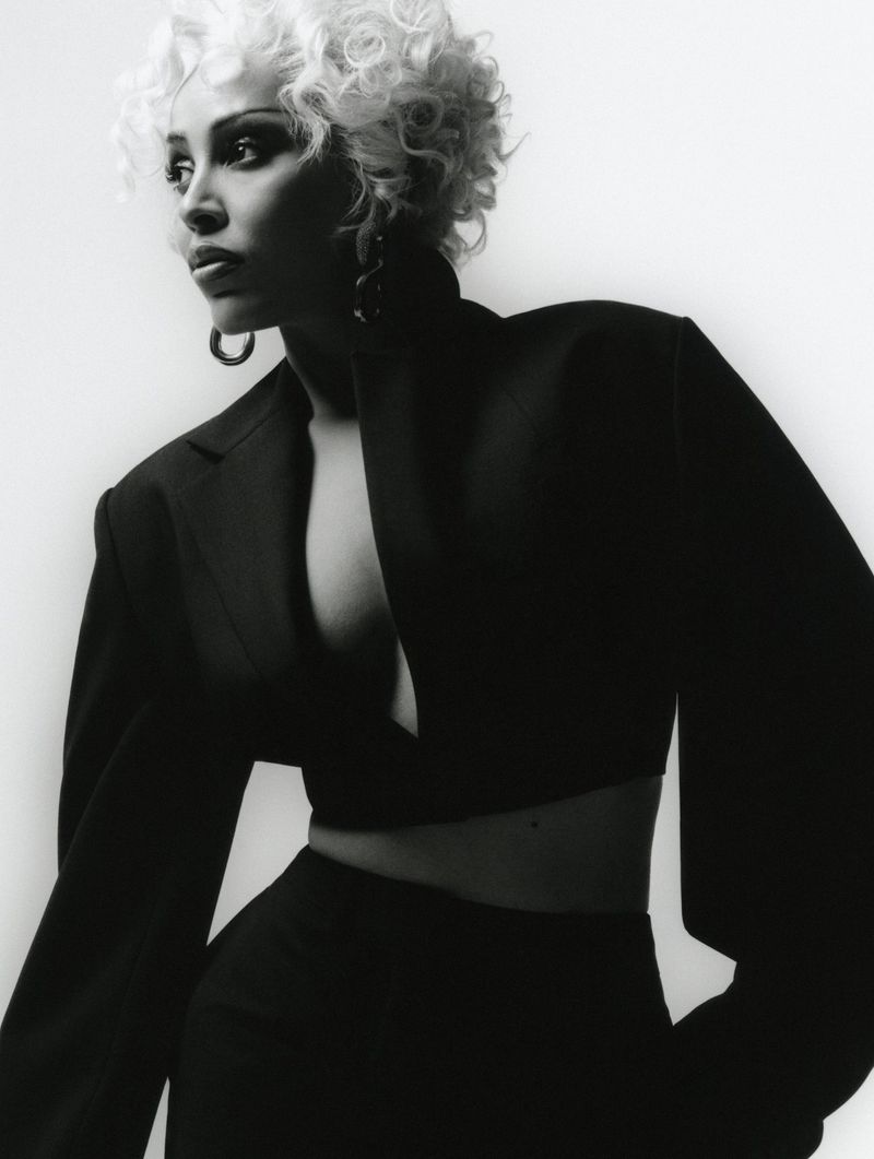 Studio shot of a young woman with short white curly hair wearing a dark crop top blazer and pants against a plain white background.