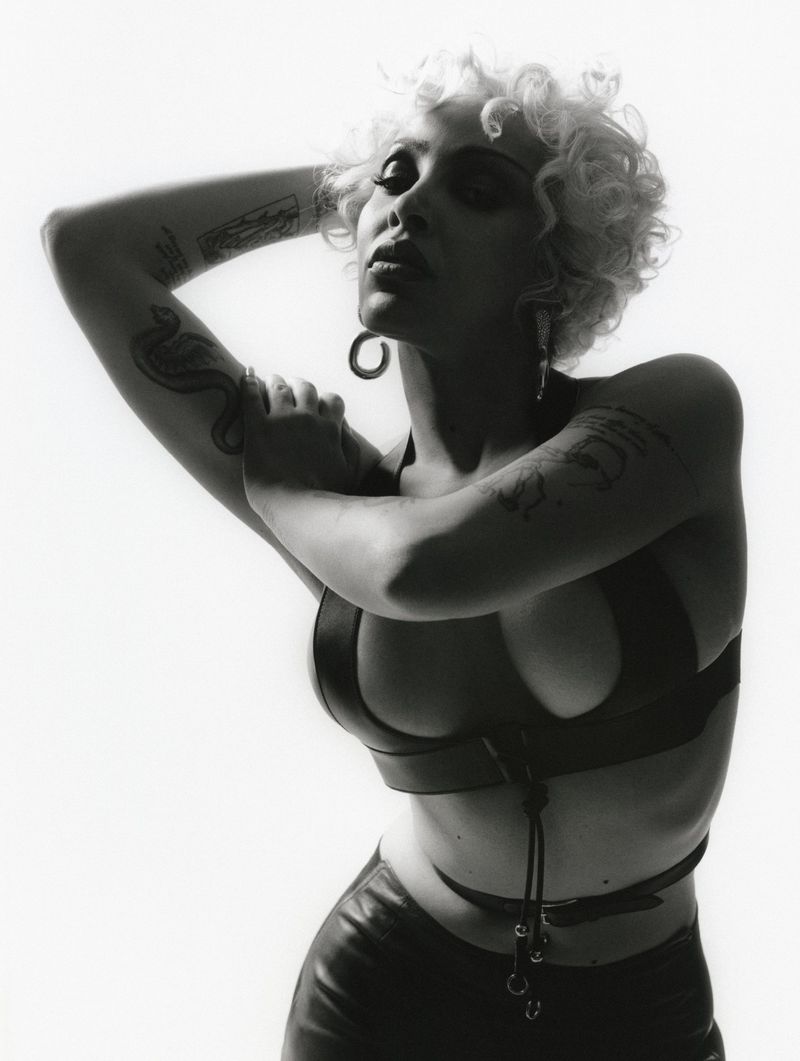 Monochrome portrait of a curly-haired woman in a modern harness-style top, arms up, gazing slightly down against a bright white backdrop.