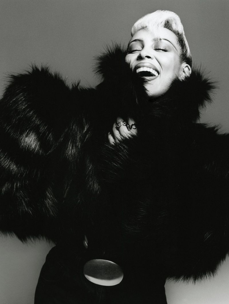 A black and white portrait of an adult woman laughing with her eyes closed, wearing a dark fur coat with a large belt buckle.