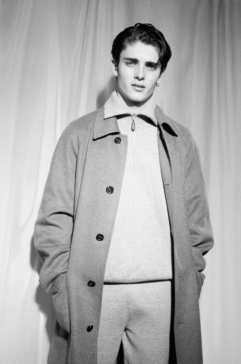 Monochrome portrait of a young adult male in a gray overcoat and matching sweater and pants, posing against a draped backdrop.