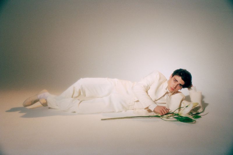A young adult male lies on his side in a white suit on a white floor next to a calla lily in a soft, evenly lit indoor studio environment.