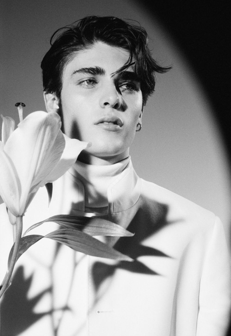 Monochrome portrait of a young adult man in a white turtleneck and blazer, holding a lily. Soft, diffused lighting creates shadows.