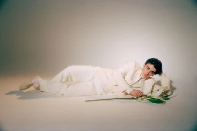 A model in a white suit reclines on the floor holding a long-stemmed white lily against a pale seamless studio backdrop.