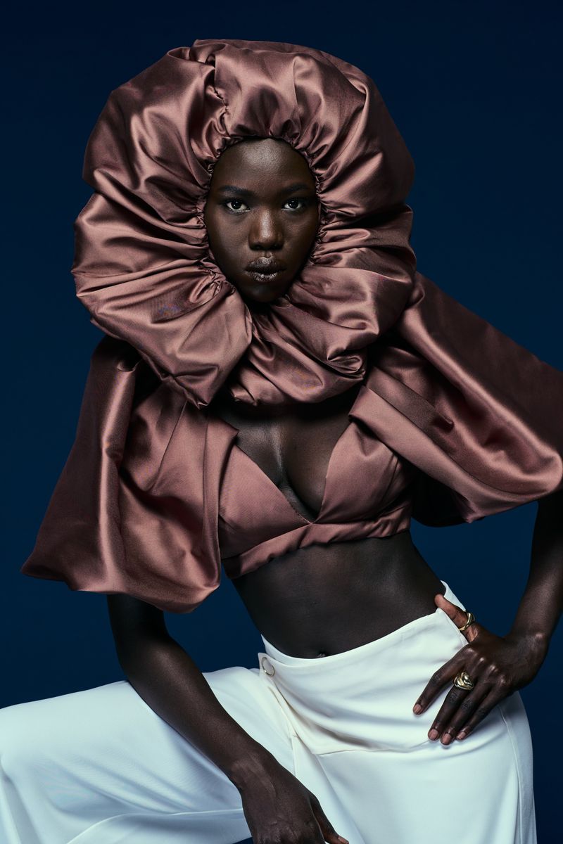 Model wearing a voluminous mauve ruffled collar and matching crop top with white trousers against a dark blue background.
