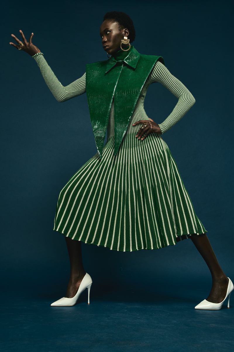 Model in a green pleated dress and glossy green vest wearing white high heels against a dark blue background