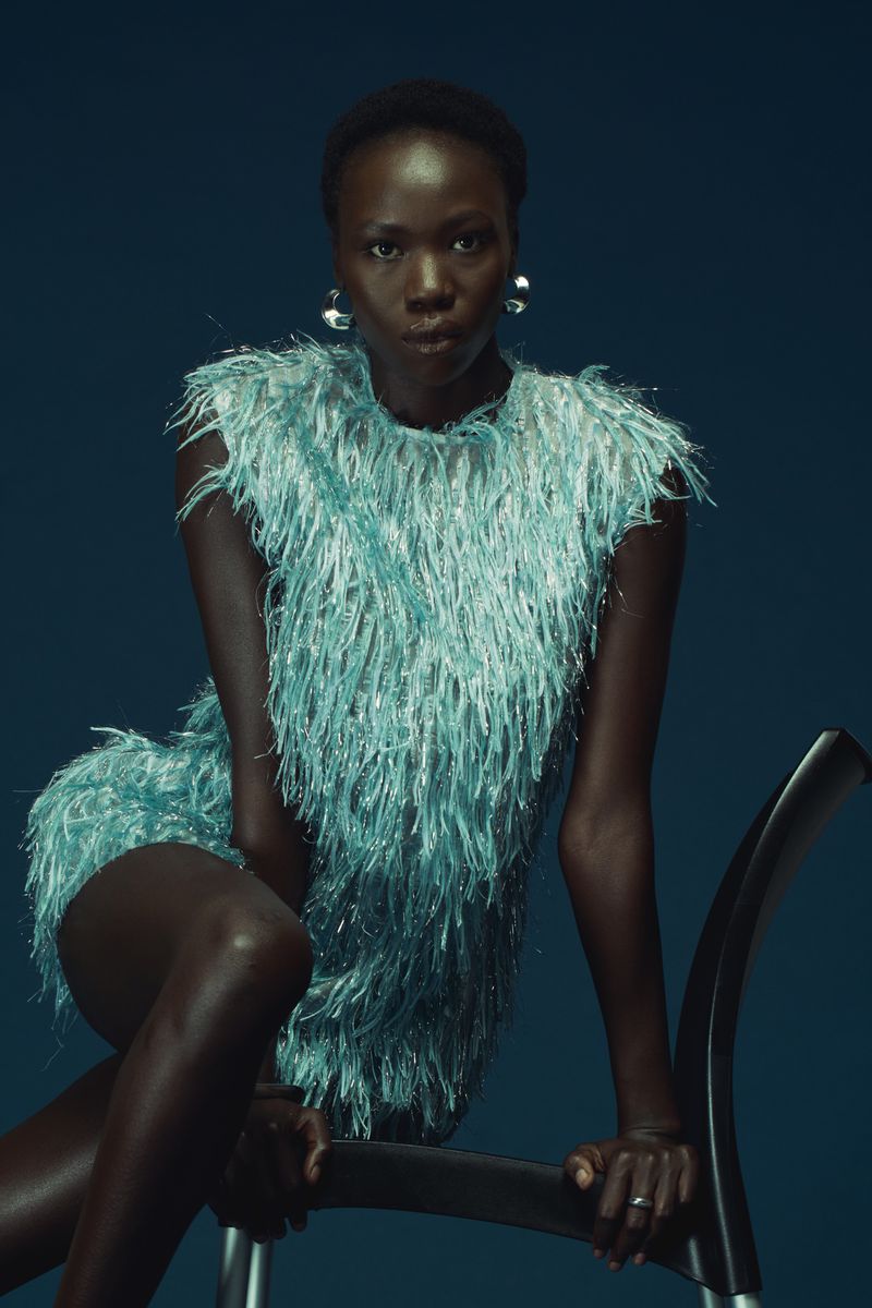 Model in a textured mint green fringed dress leaning over a dark curved chair against a dark blue background.