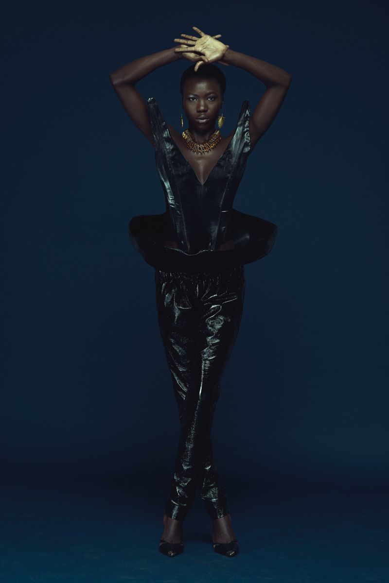 Black woman with gold hands raised above head wearing structured black top and trousers against dark blue studio backdrop.