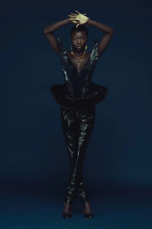 Black woman with gold hands raised above head wearing structured black top and trousers against dark blue studio backdrop.