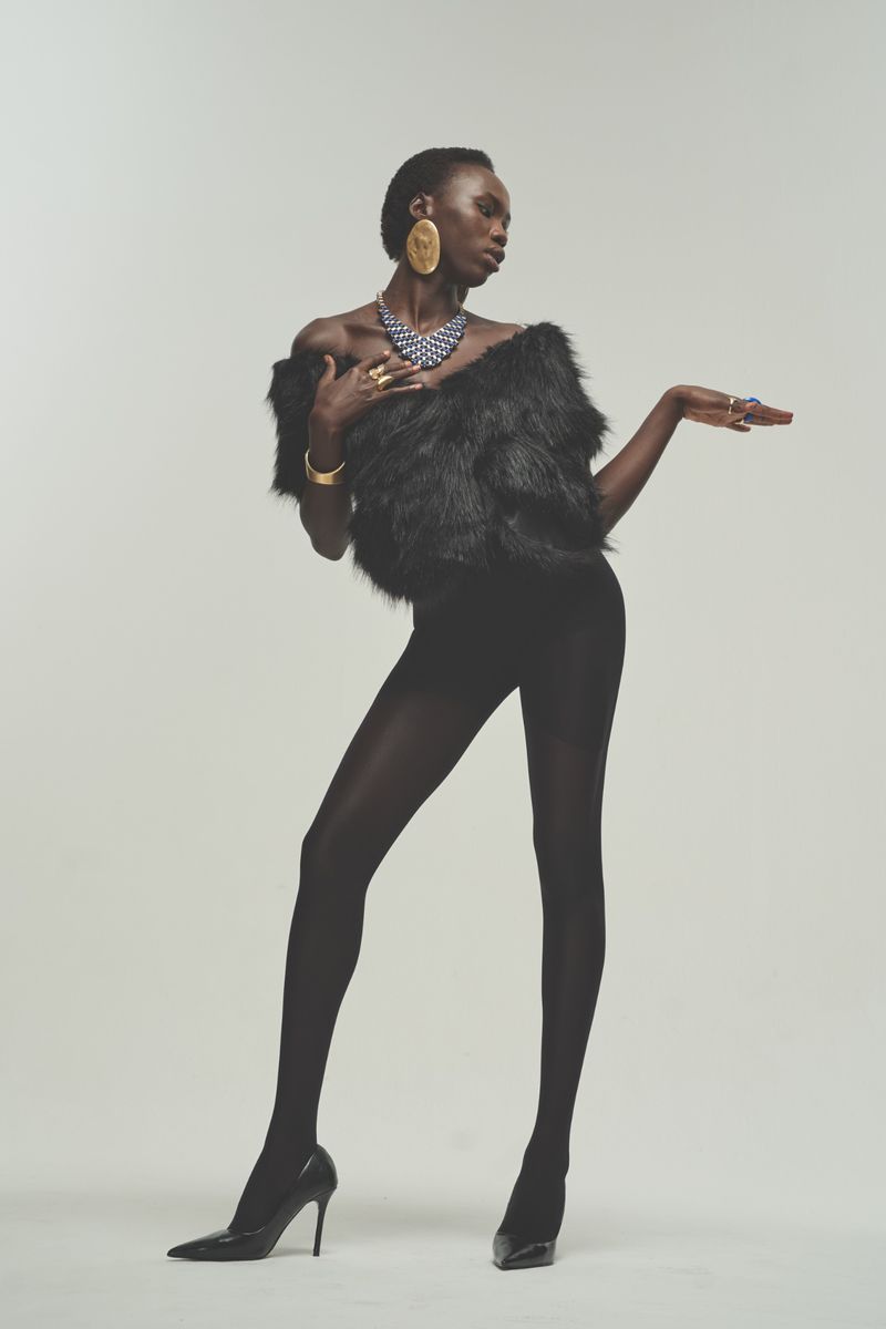 Model in black textured top and black tights wearing gold jewelry against a plain light background for Glamour Magazine U.S.