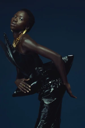 Black woman wearing a glossy black sculpted garment with large gold earrings against a dark blue background.