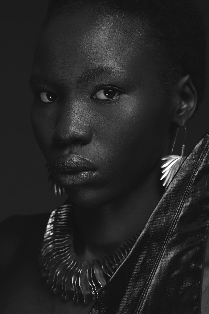 Close-up portrait of a Black woman wearing statement silver earrings and a large textured necklace against a dark background.