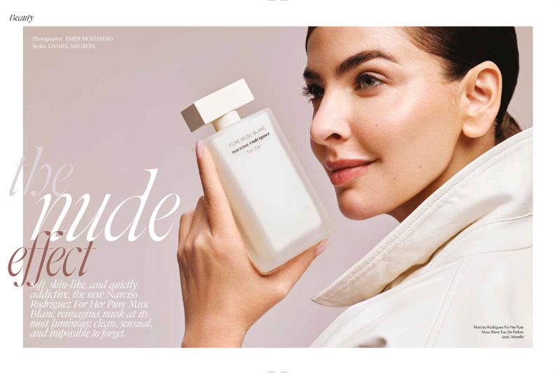 Close-up of a woman holding a white perfume bottle near her face, wearing a cream coat against a light background.