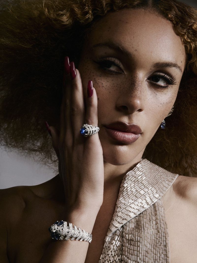 Close-up portrait of a woman wearing diamond and sapphire jewelry, a beaded dress, and holding her hand to her face with dark red nails