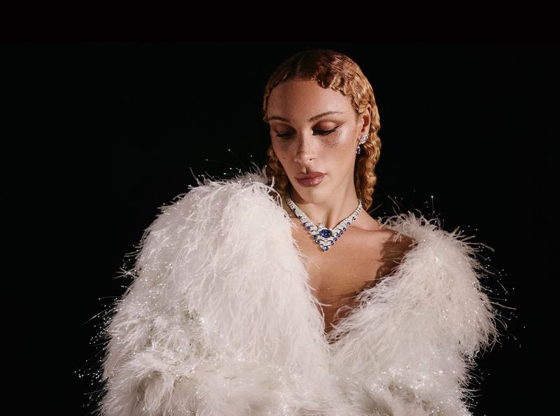 Model wearing a white feathered coat and jeweled necklace against a dark background for the La Gema project