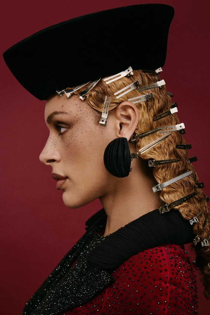 Profile portrait of a woman wearing a black hat, metal hair clips, statement earring, and a red textured top against a burgundy backdrop.