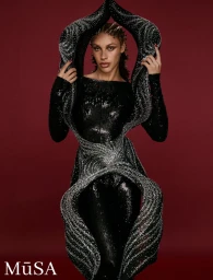 Model wearing a sculptural black sequined garment with intricate silver embellishments against a dark red background.