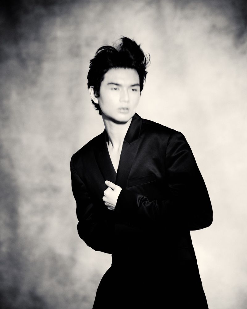 Monochrome portrait of a stylish Asian male model wearing a black suit jacket against a textured light-gray background.