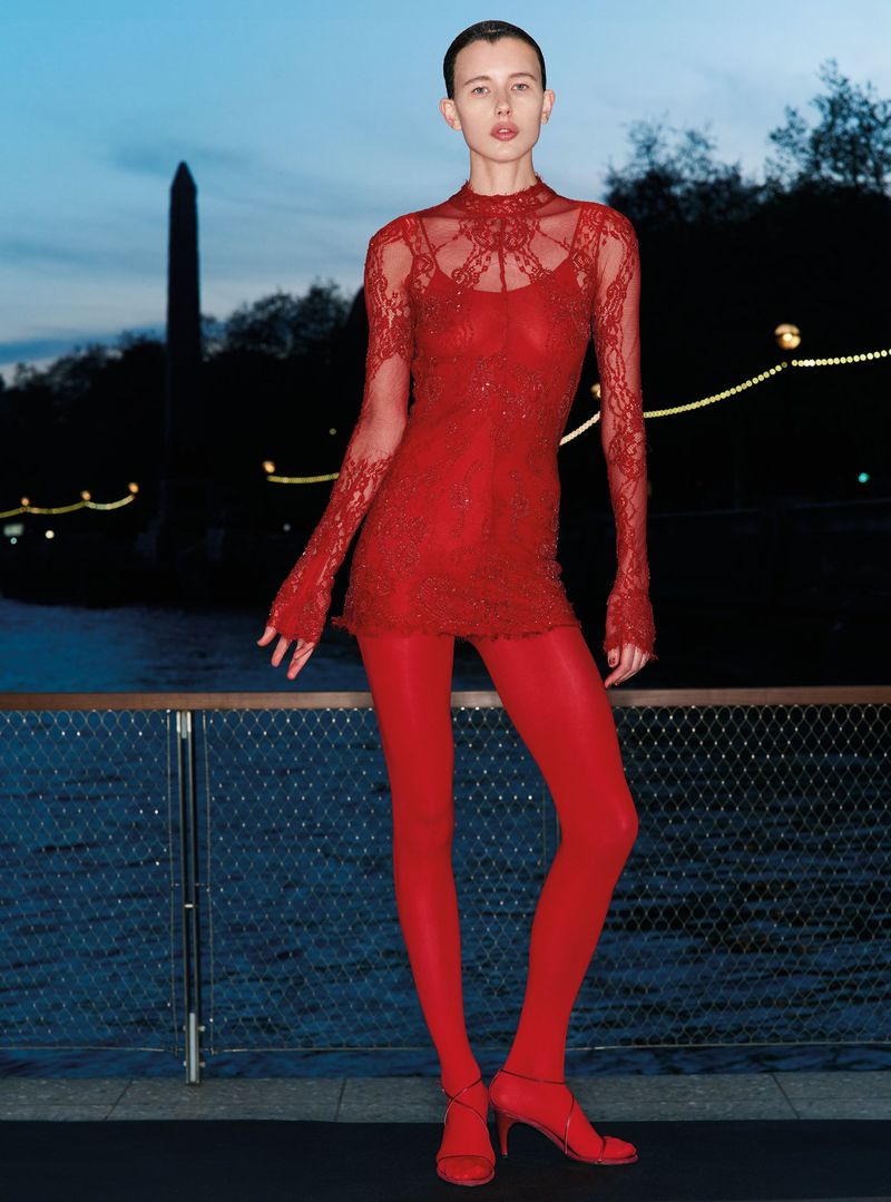 Model in a red lace dress and matching red tights standing against an evening waterfront with an obelisk silhouette.